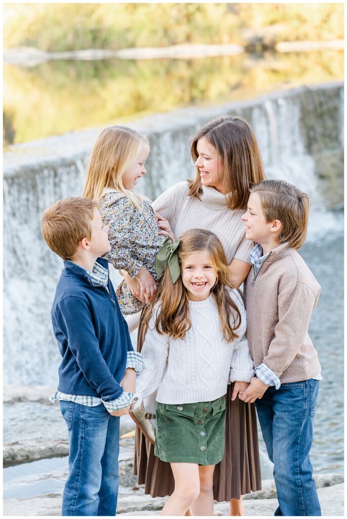 Mother and her two boys and two girls standing in front of a waterfall at blue hole park. She holds and tickles her youngest daughter while the other children laugh in this candid shot captured by a Georgetown Texas Photographer