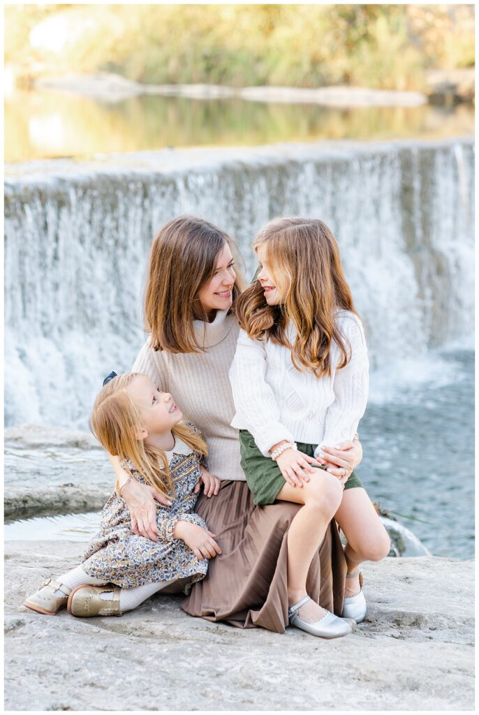 A mother and her two young daughters sit in front of a waterfall at blue hole park. The mother and youngest daughter look at the older daughter. They all smile together in this sweet moment captured by a Georgetown Texas Photographer.