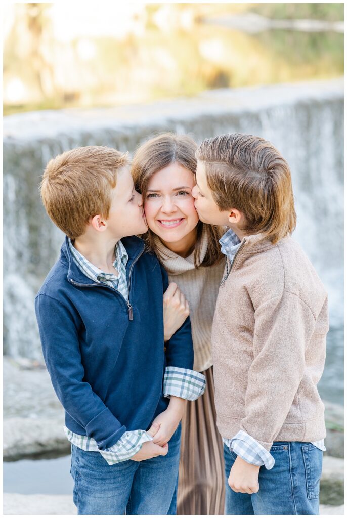 A mother and her two young sons stand in front of a waterfall in Georgetown Texas. The two boys kiss their mother on the cheeks in this sweet photo taken by a Georgetown Texas Photographer.