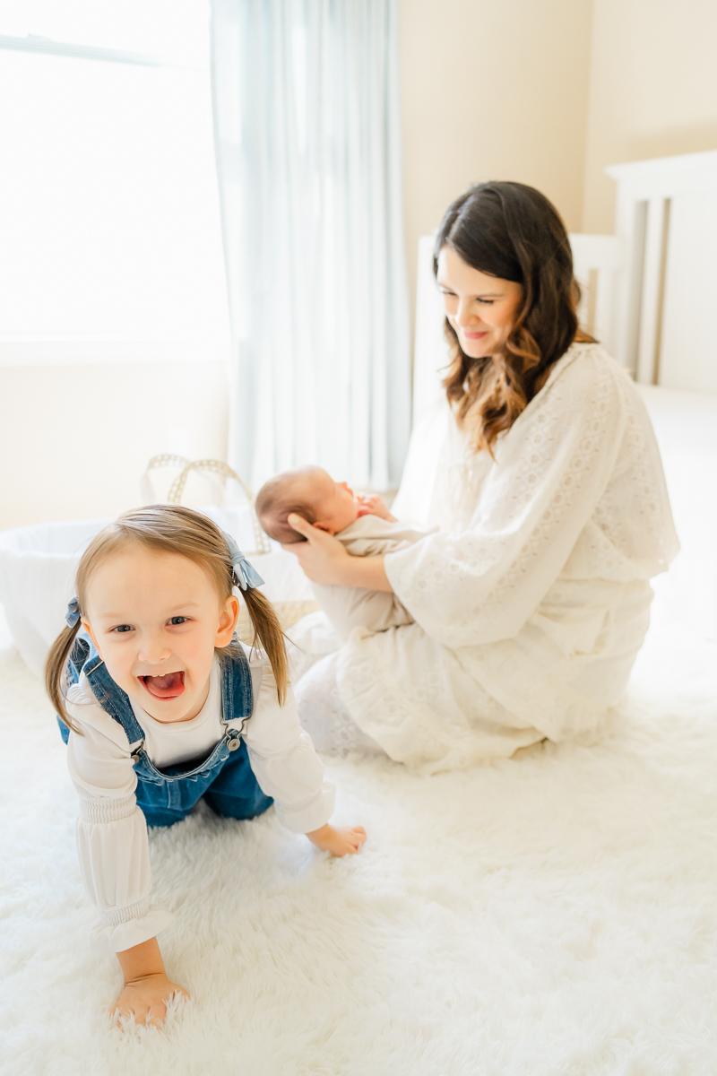 a mother looks down at her young daughter while holding her newborn son. They are in a bright nursery, captured in a candid moment by an Austin Photographer.