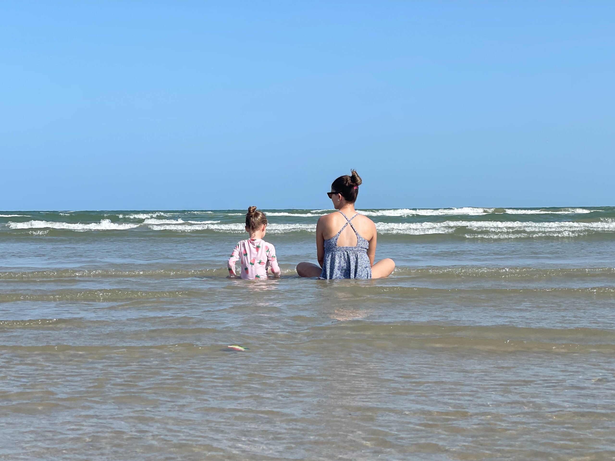 a mother and daughter sit in the water at the beach on the Texas Gulf Coast. They look at the horizon together in this sweet photo taken by an Austin Photographer.