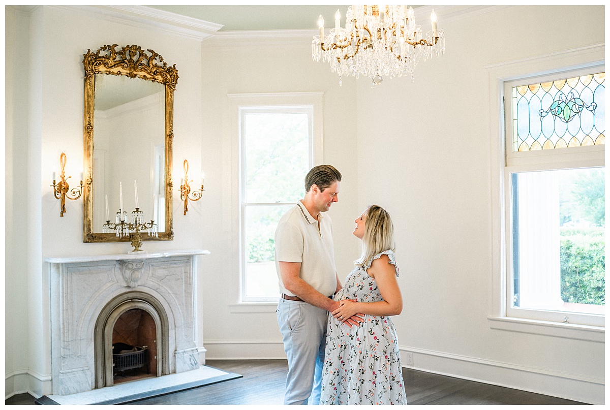 A husband and wife stand facing each other under a chandelier inside Woodbine Mansion in Round Rock, TX. They look at each other smiling while holding their hands on the wife's pregnant belly in this tender photo taken by an Austin Maternity Photographer.