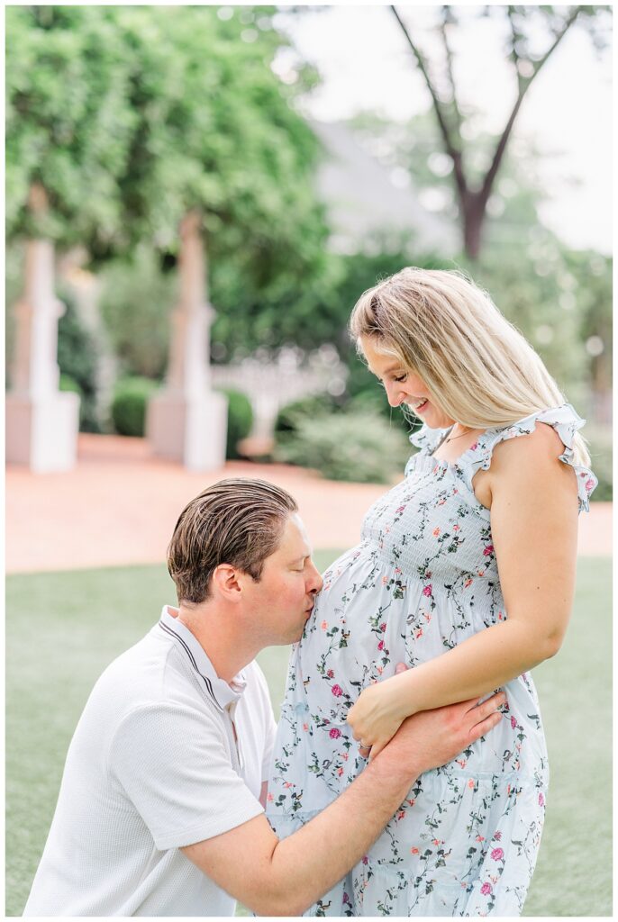 A husband is on his knees and kisses his wife's pregnant belly. She looks down at with a smile on her face.