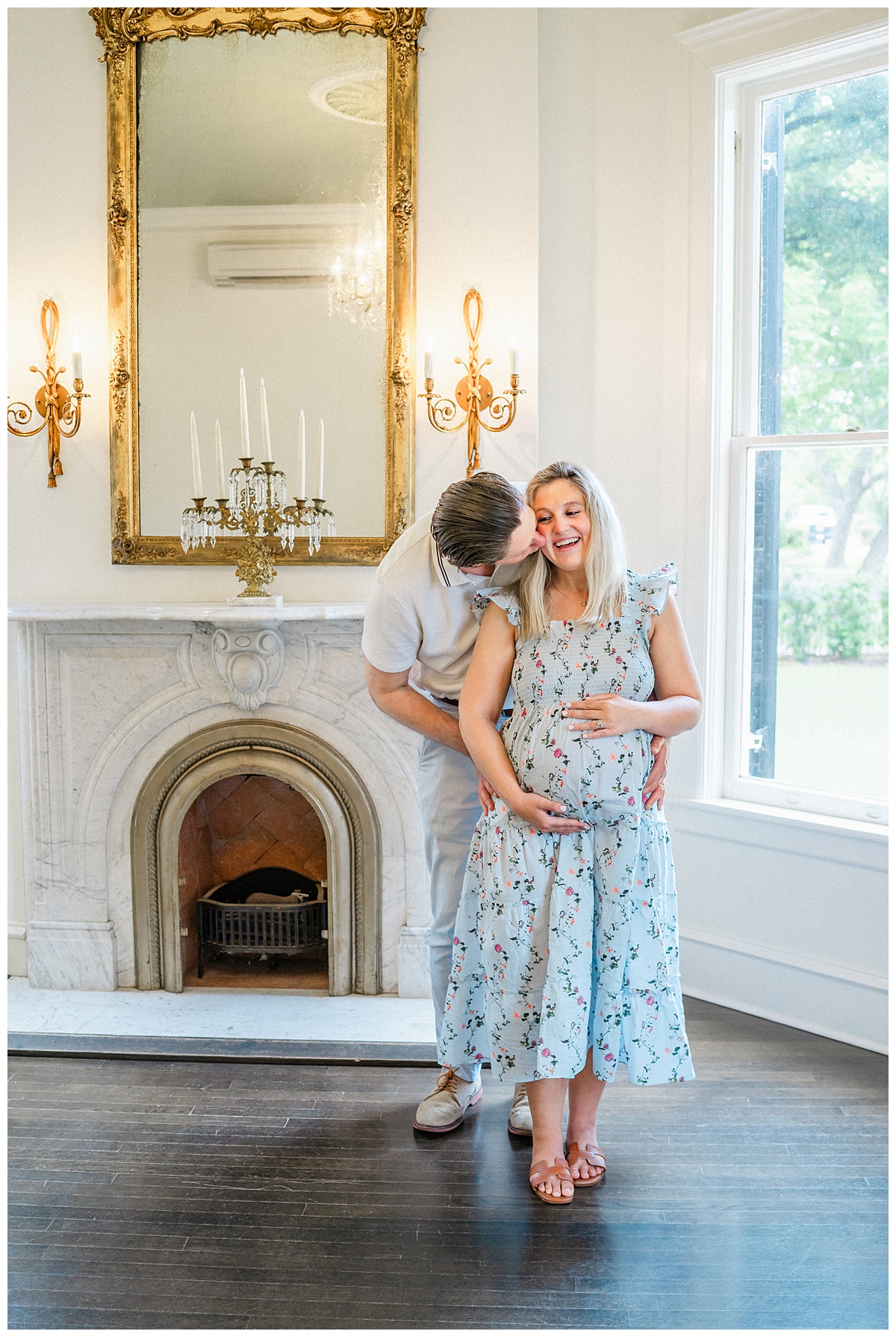 her husband and his pregnant wife stand in front of a fireplace at the Woodbine Mansion. The husband kisses his wife on the cheek while holding her belly, beautifully captured by an Austin Maternity Photographer.