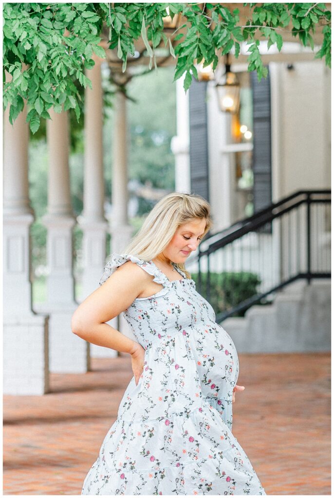 A pregnant mother stands under the white columns at the Woodbine Mansion. She is dressed in a blue floral dress and looks down at her belly with a soft smile on her face.