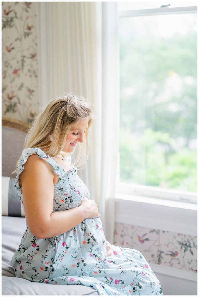 A pregnant mother sits on a mauve daybed at the Woodbine Mansion. Beautiful natural light is coming through the window and beautifully reflecting on her skin. She smiles and looks down at her belly.