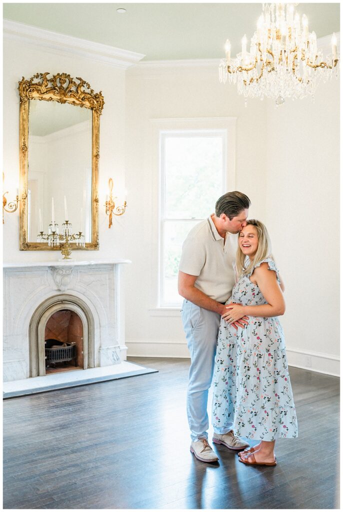A husband and wife stand under a chandelier and in front of a fireplace at the Woodbine Mansion. The husband kissed his pregnant wife on the forehead while she closes her eyes and smiles in this sweet photo taken by an Austin Maternity Photographer.