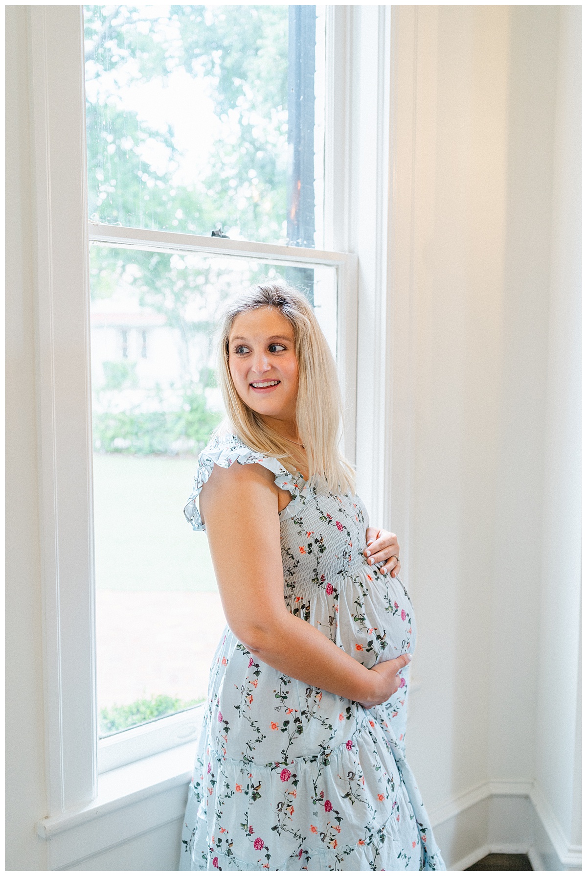 A pregnant woman with blond hair and dressed in a floral midi dress stands in front of a window at woodbine mansion. She looks away while placing her hands on her belly, in this shot captured by an Austin Maternity Photographer.