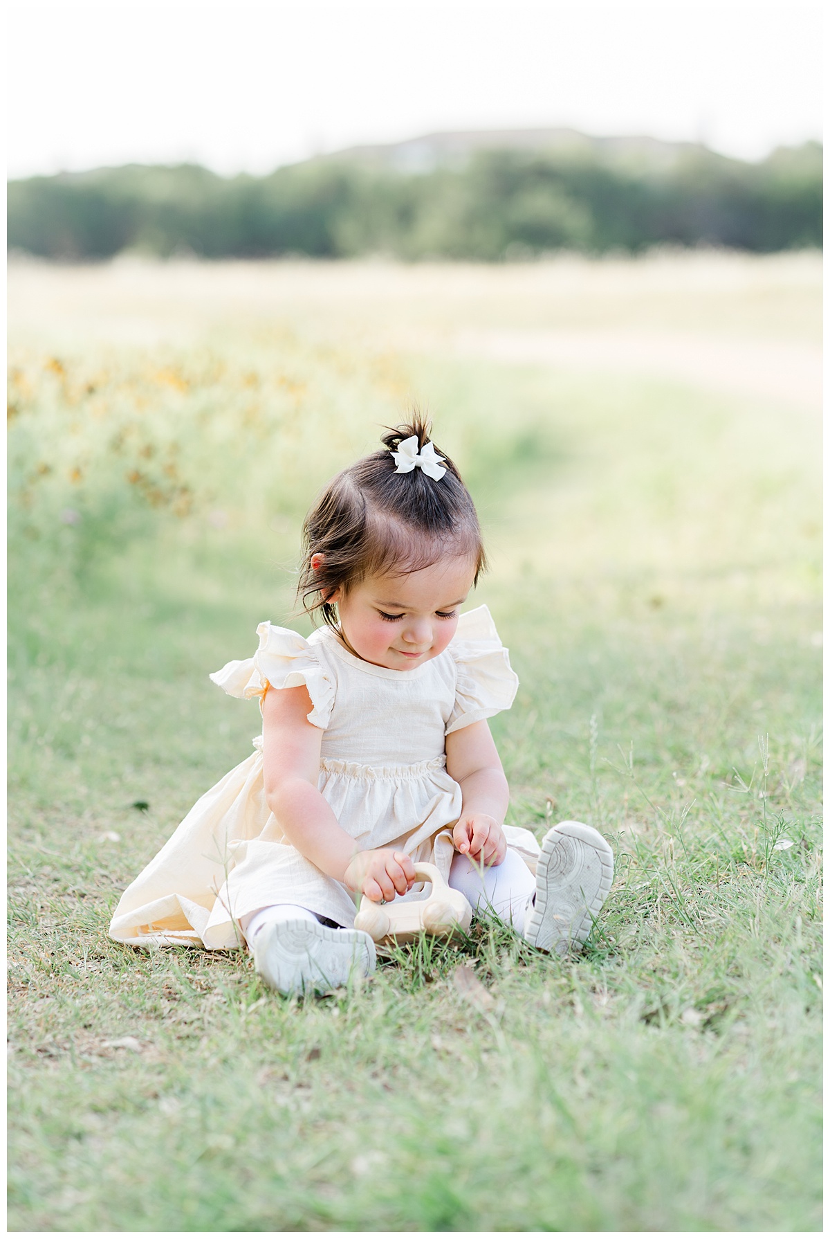 A toddler girl dressed in a soft yellow dress and white tights sits in grass in front of a field of wildflowers. She looks down and smiles at a toy car she plays with during her wildflower family photos in Cedar Park, TX