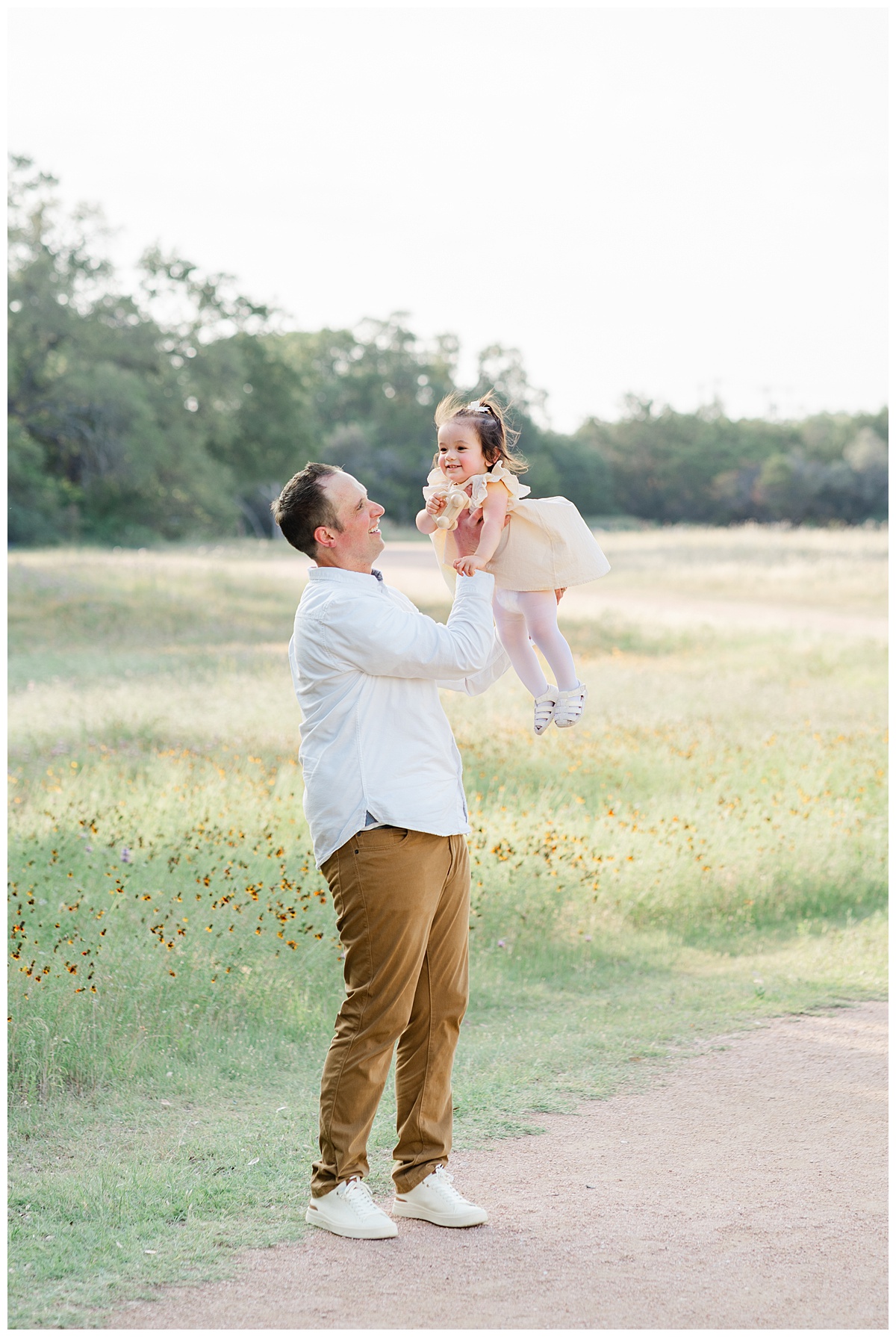 A father stands in front of a field of wildflowers and throws his toddler girl in the air. The little girl smiles in this candid moment taken during wildflower family photos.