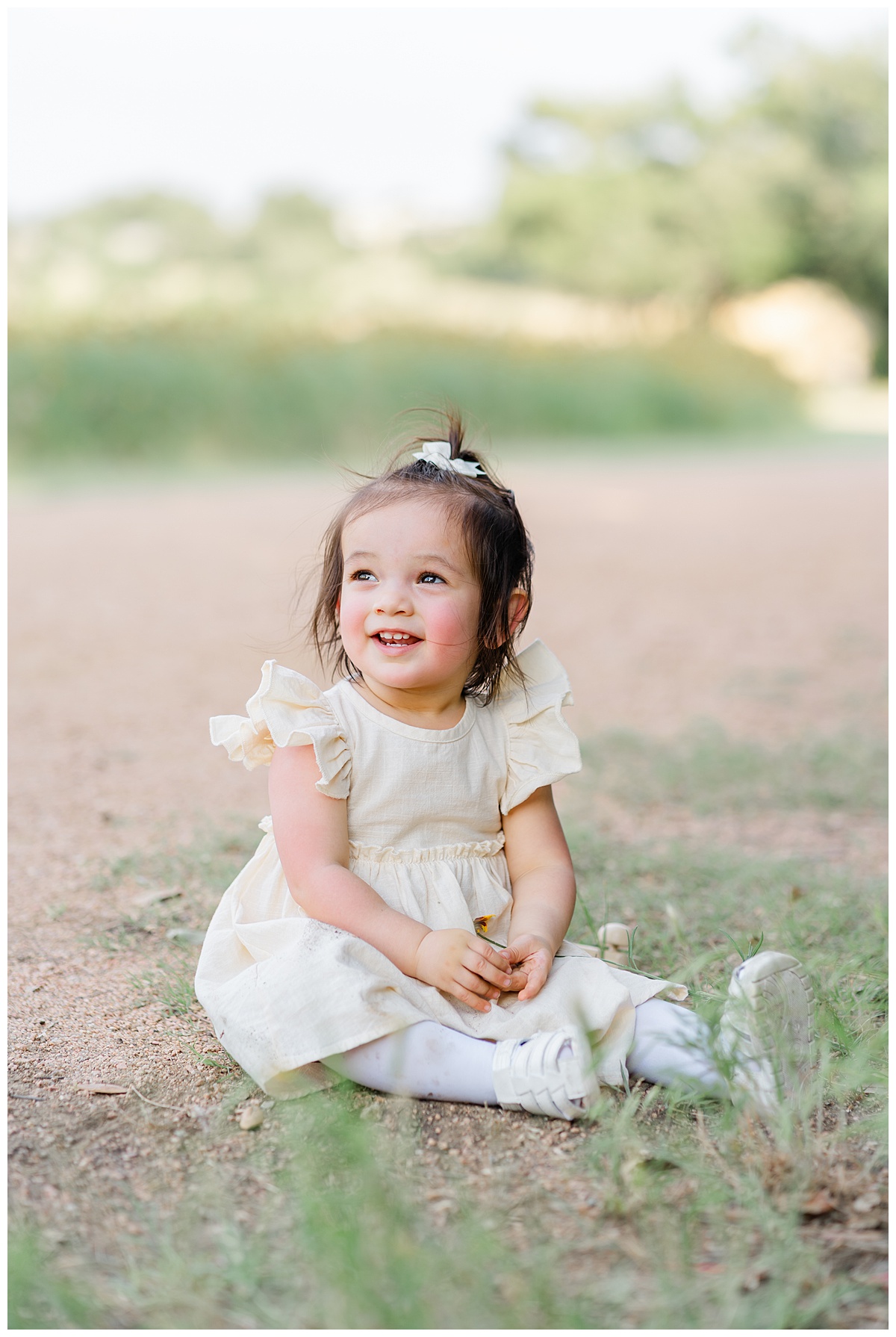 A toddler girl sits on a sandy path in Cedar Park, TX. She looks up and smiles in this sweet photo taken by a Cedar Park Family Photographer.