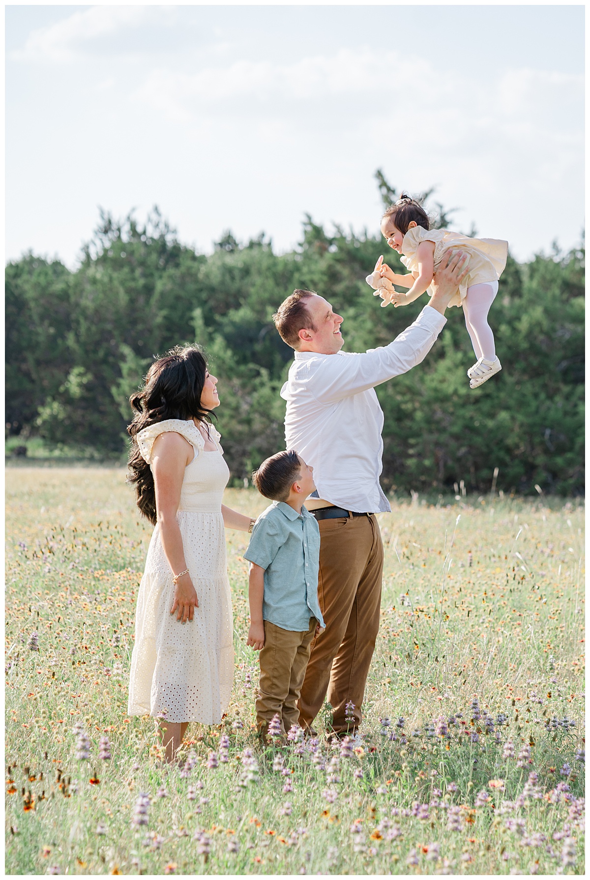 A family of 4 stands in a field of wildflowers for their family photos in Cedar Park, TX. The father throws their little girl in the air while the mom and big brother look at her smiling.