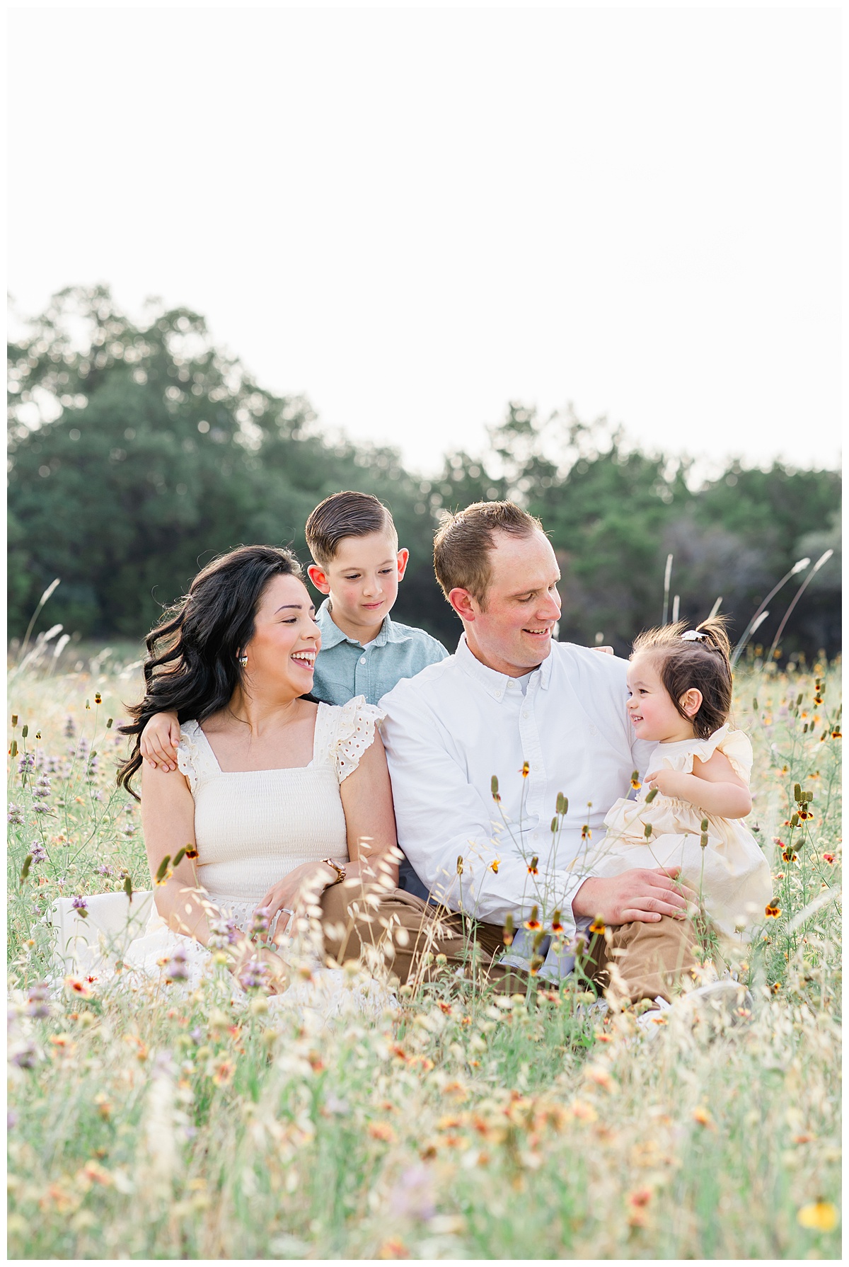 family of four sits in a field of wildflowers and laughs with each other, candidly captured by a Cedar Park Family Photographer