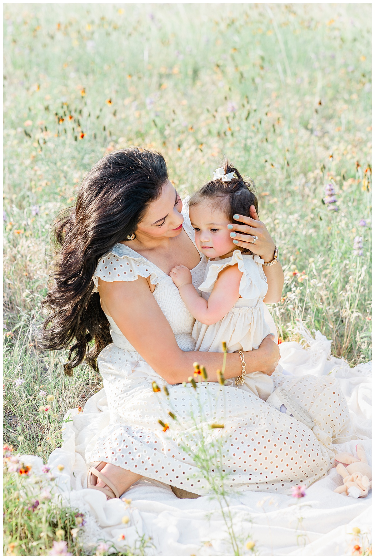 A mother sits on a white blanket in a field of wildflowers. She hold her toddler girl on her lap and looks down at her in this tender moment captured by a Cedar Park Family Photographer.