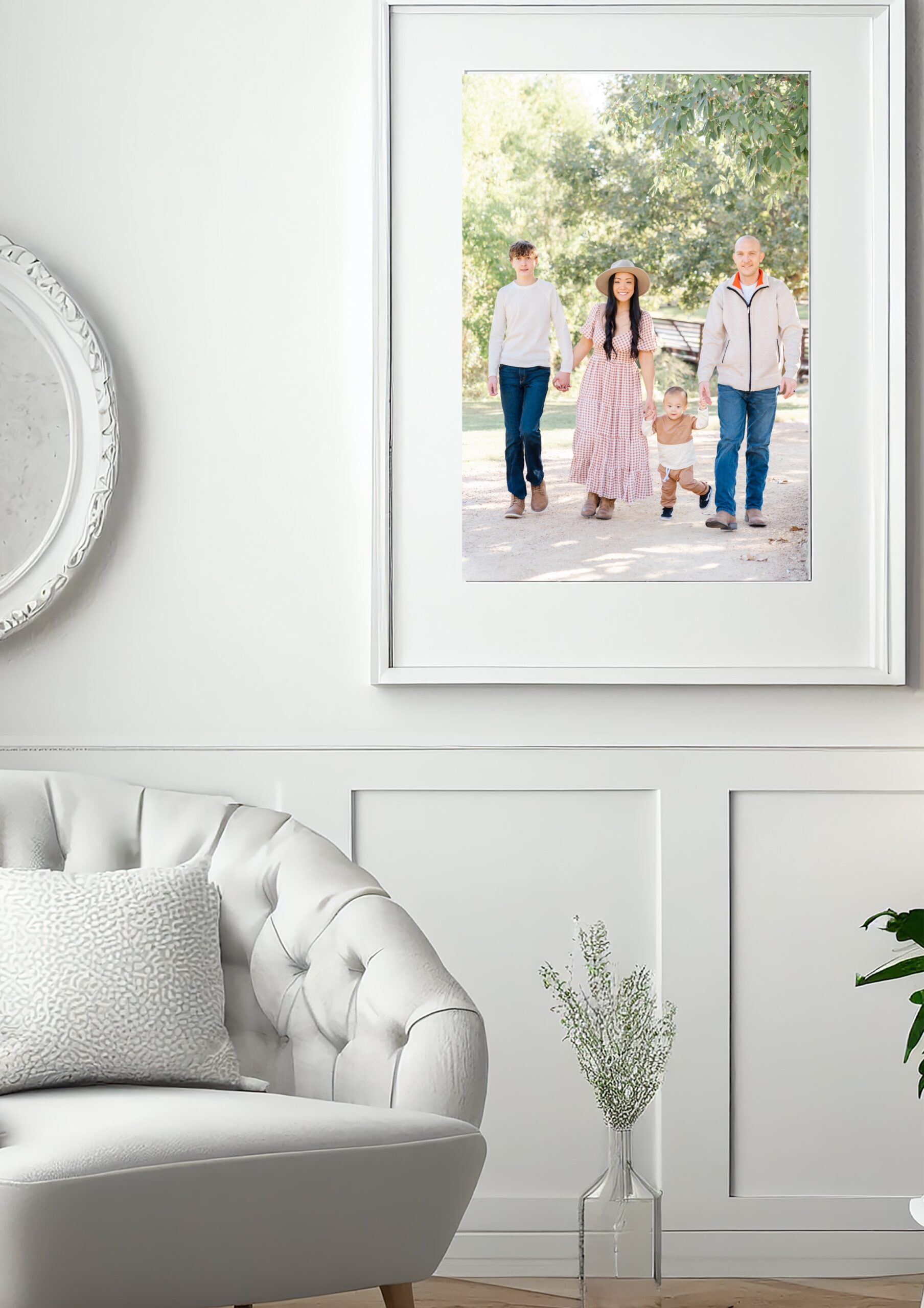 a picture of a large framed portrait on a white wall with wainscotting. In the corner is an el.egant white chair. The portrait is of a family of four walking up a sandy path at a park.