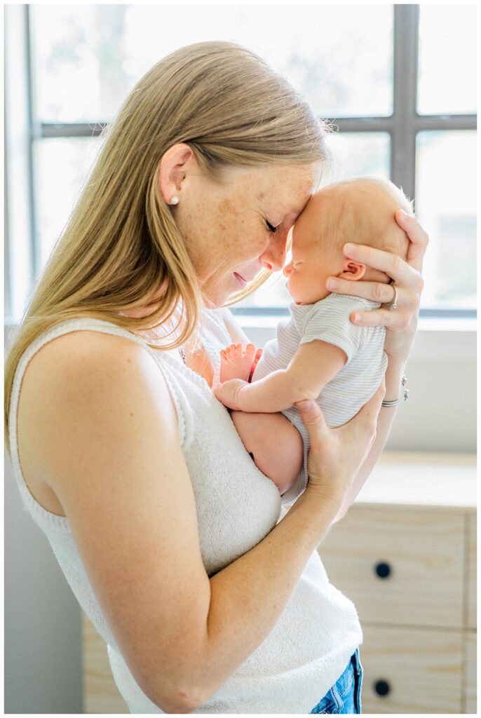 a mother stands in front of a window in her Georgetown, TX home while holding her newborn baby facing her and resting her head against him.