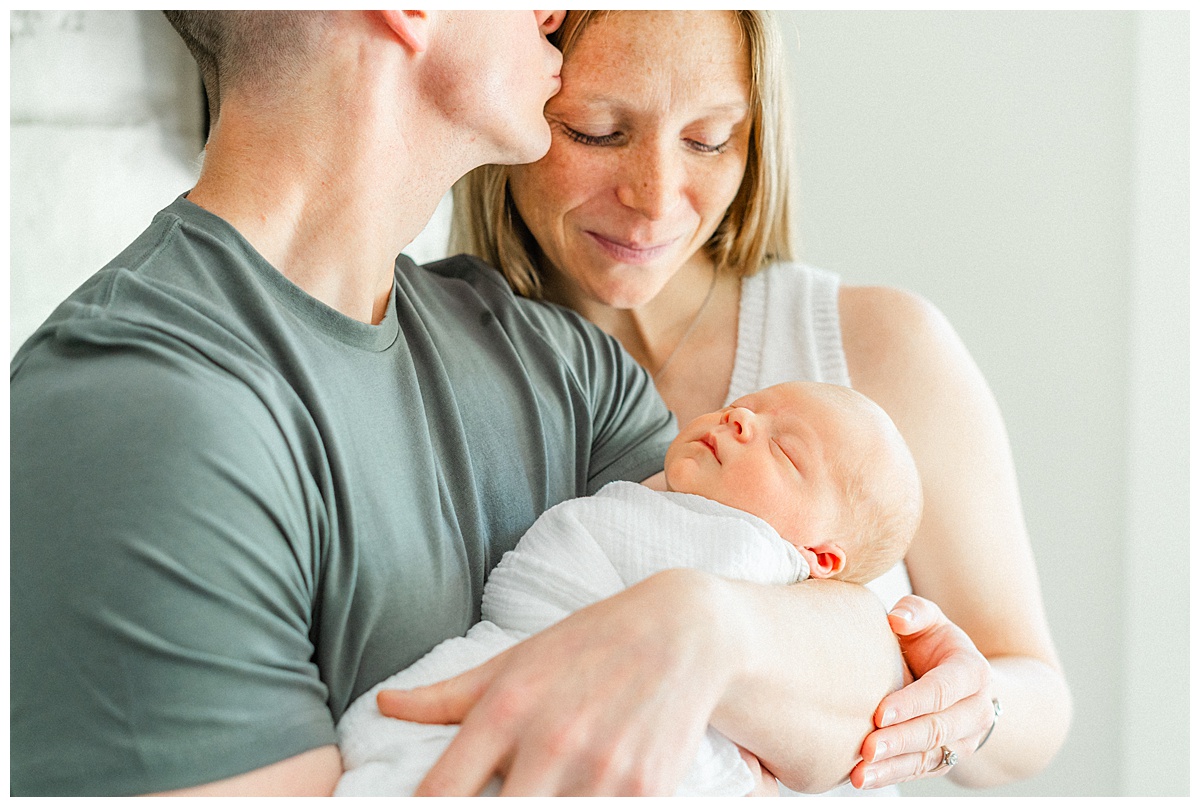 A father holds his newborn baby while kissing his wife on the temple in this photo captured by an in-home newborn photographer in Georgetown, TX