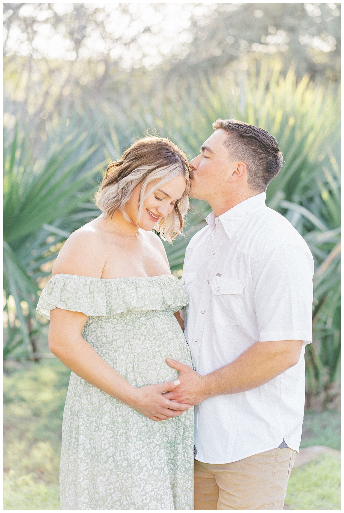 A pregnant wife and her husband stand in front of a palm tree at Bull Creek District Park. The husband kisses his wife on the temple during their maternity photoshoot in Austin, TX.