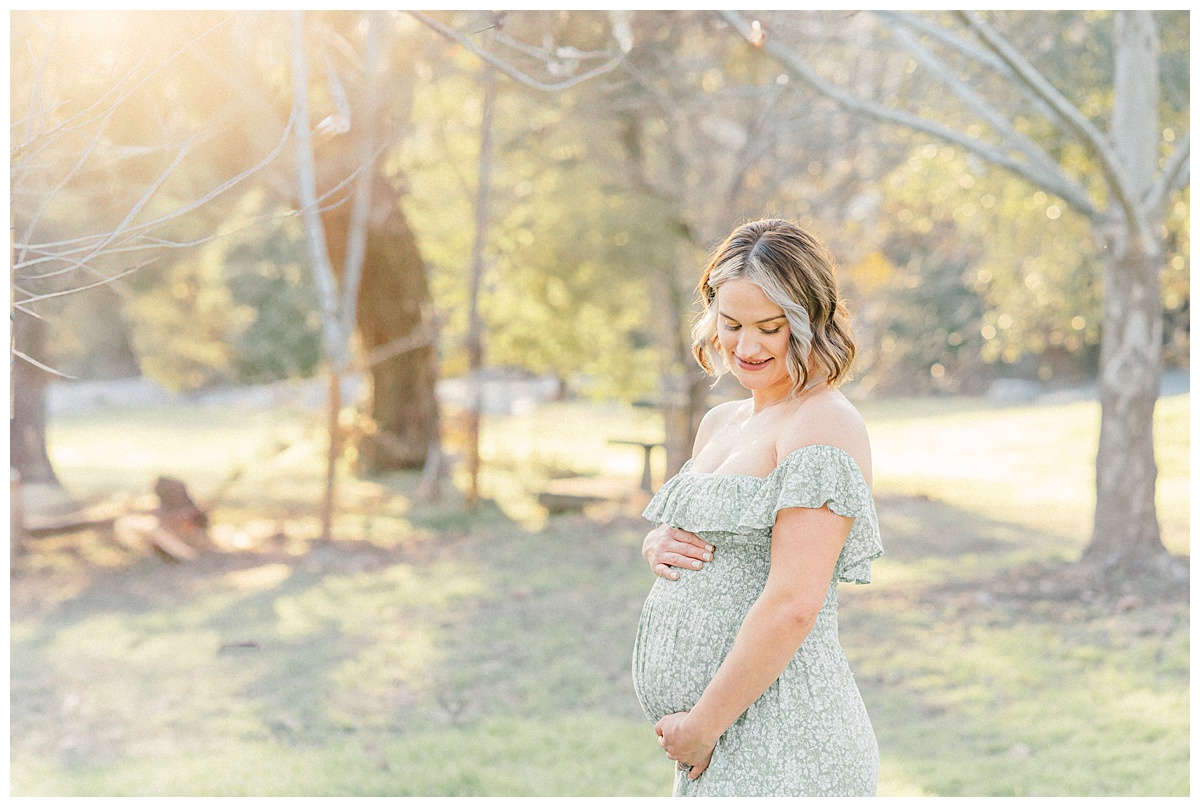 A pregnant mother, captured by an Austin Maternity Photographer, stands under a tree at Bull Creek District Park. She holds her belly and looks down with a soft smile.
