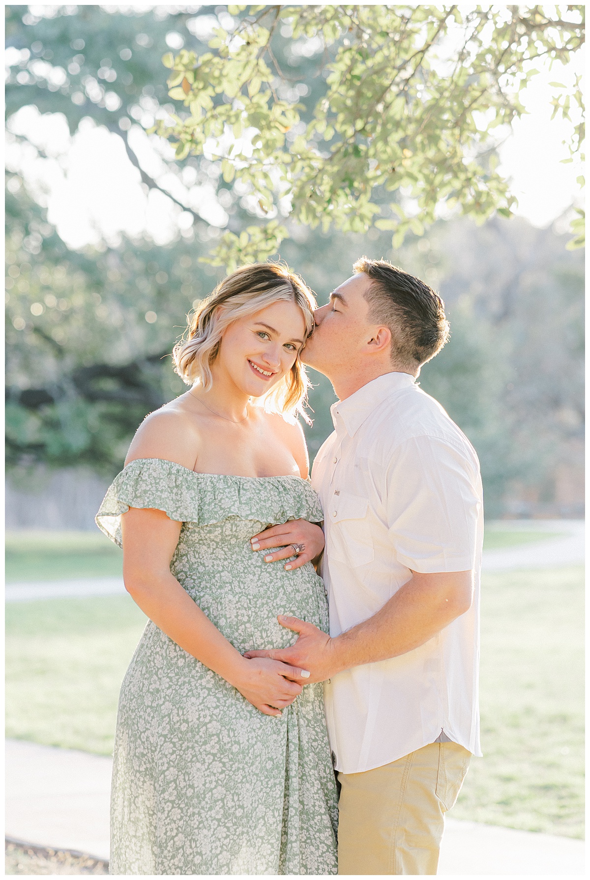 A couple stands under a tree at Bull Creek District Park. The pregnant wife looks at the camera of her Austin Maternity Photographer and smiles. Her Husband kisses her on the temple with his hand on her belly.