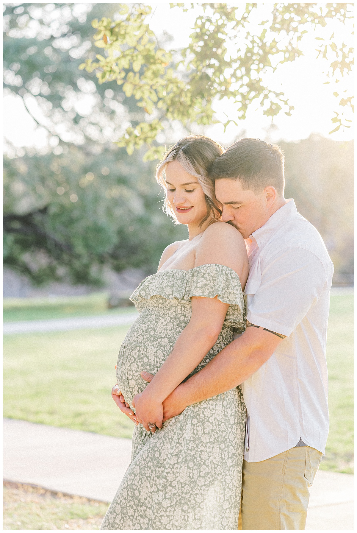 A couple stands under a tree at Bull Creek District Park. The husband stands behind his pregnant wife and kisses her shoulder. They both look down at her belly in this shot captured by an Austin Maternity Photographer.