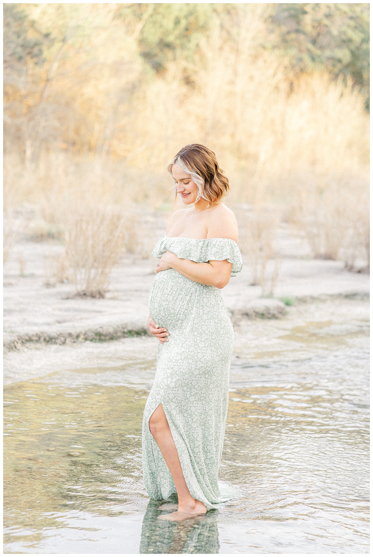 A pregnant mother stands in the water at Bull Creek District Park. She places her hands on her belly and looks down at it. She smiles, in this tender shot captured by an Austin maternity photographer.