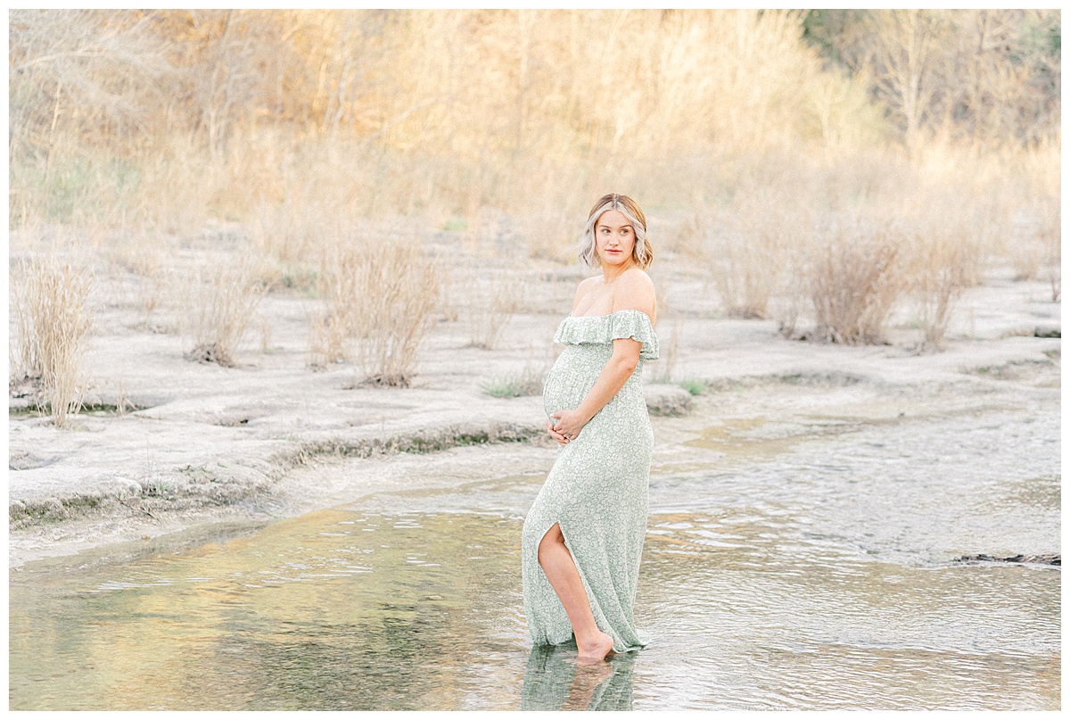 a pregnant mother stands in the water at Bull Creek District Park. Dressed in a long green dress, she holds her belly with both hands while looking away during her maternity photoshoot in Austin, TX.