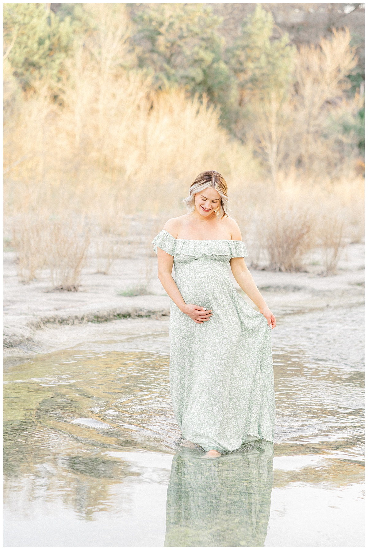 A pregnant woman dressed in a long green dress walks in the water at Bull Creek District Park. She looks down at the water while holding her belly in this shot captured by an Austin Maternity Photographer.