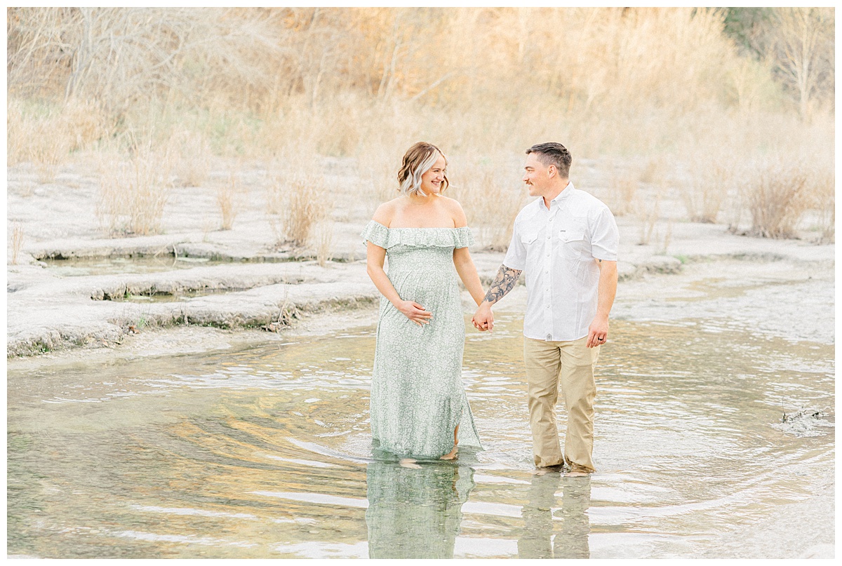 A couple walks in the water at Bull Creek District Park while holding hands and looking at each other. The woman, dressed in a long green dress, holds her belly with one hand during her maternity photoshoot in Austin, TX.