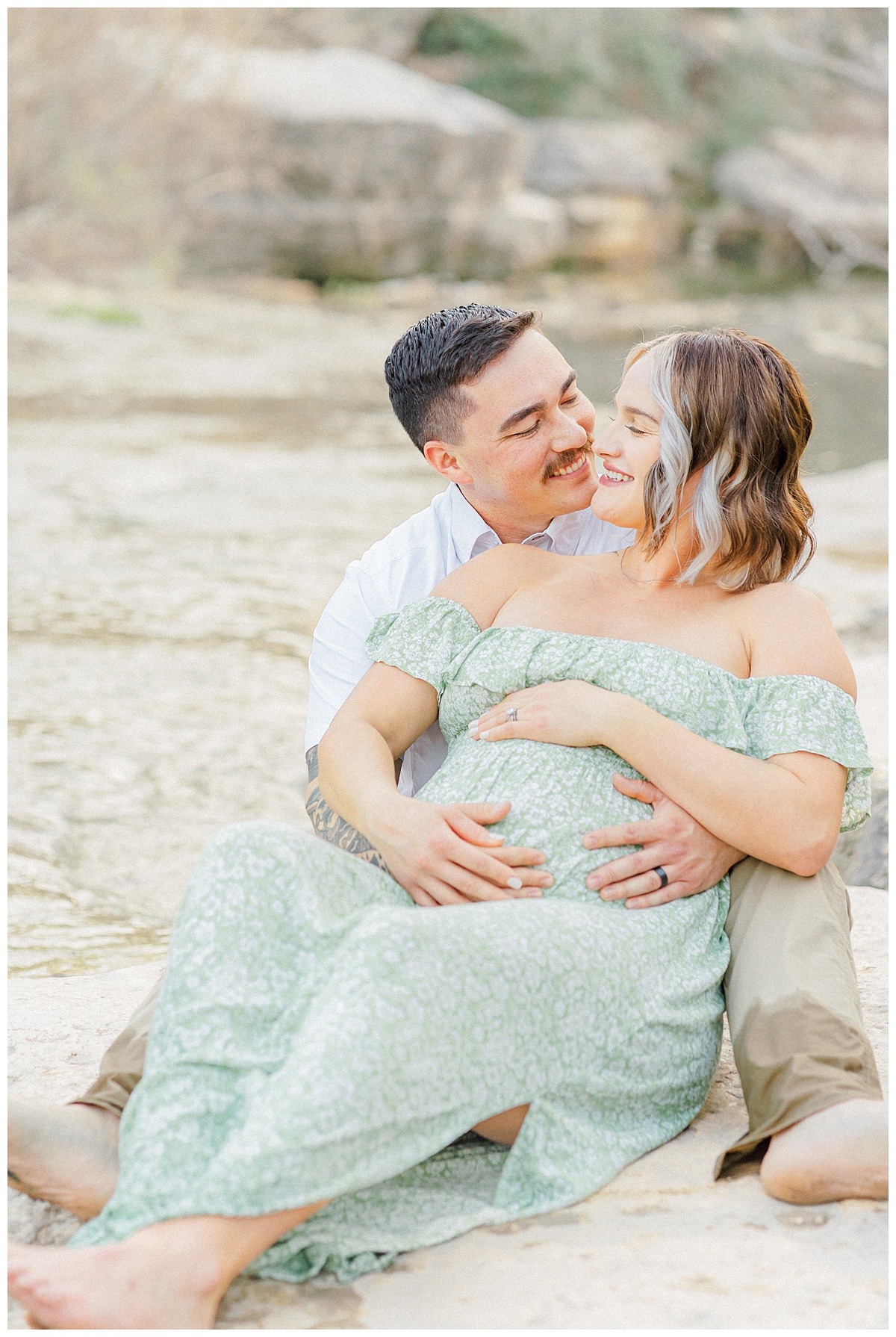 A couple sits by the water at Bull Creek District Park. The pregnant woman leans against her husband. They close their eyes and smile while being captured by an Austin maternity photographer.