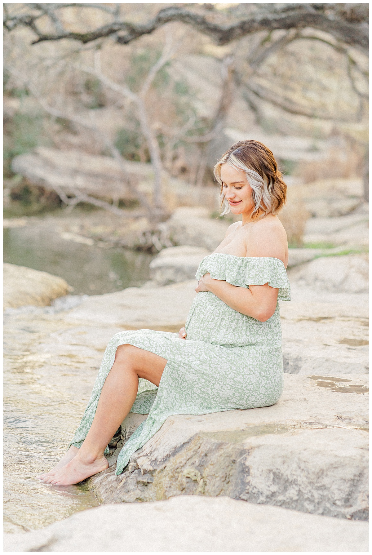 A pregnant woman sits by the water at Bull Creek District Park. Dressed in a long green dress, she holds her belly with both hands and looks down at her belly with a smile during her maternity photoshoot in Austin, TX.