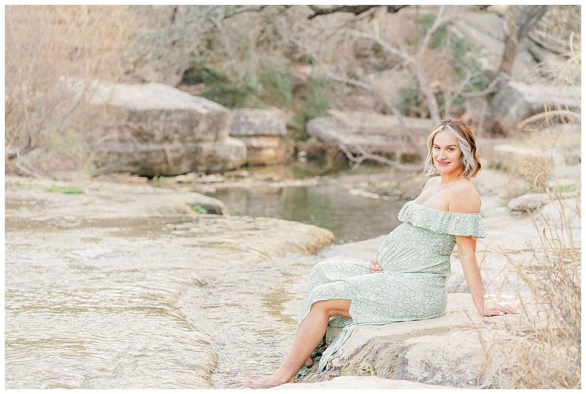 A pregnant woman sits by the water at Bull Creek District Park. She is dressed in a long green dress. She holds her belly while looking at the camera and smiling.