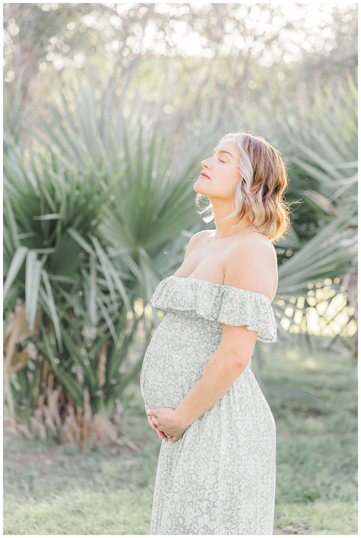 A pregnant mother stands in front of a palm tree at Bull Creek District Park. She closes her eyes while holding her belly in this sweet photo captured by an Austin Maternity Photographer.