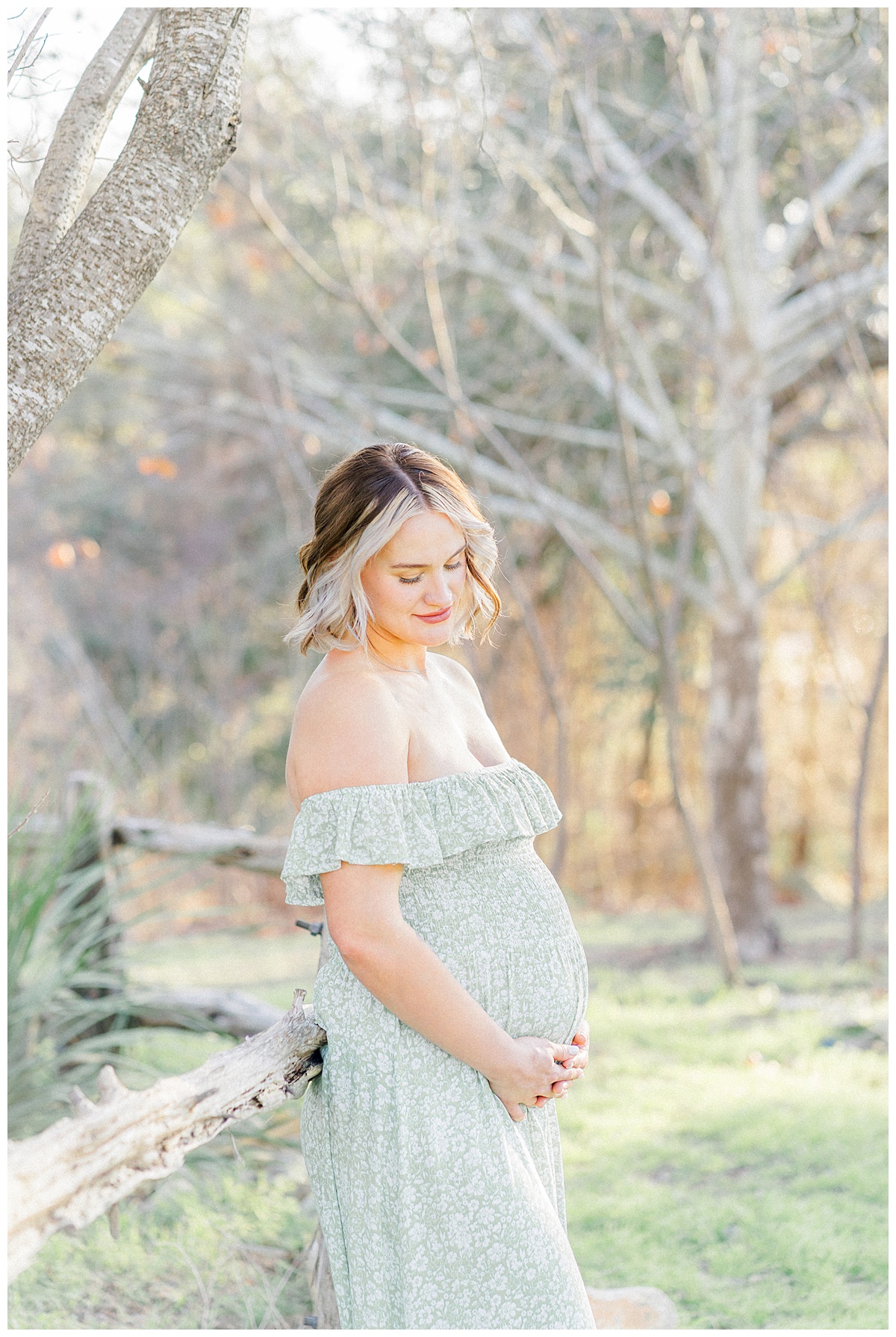 A pregnant woman leans against a rustic fence at Bull Creek District Park. Dressed in a long green dress, she looks down at her belly with a soft smile.