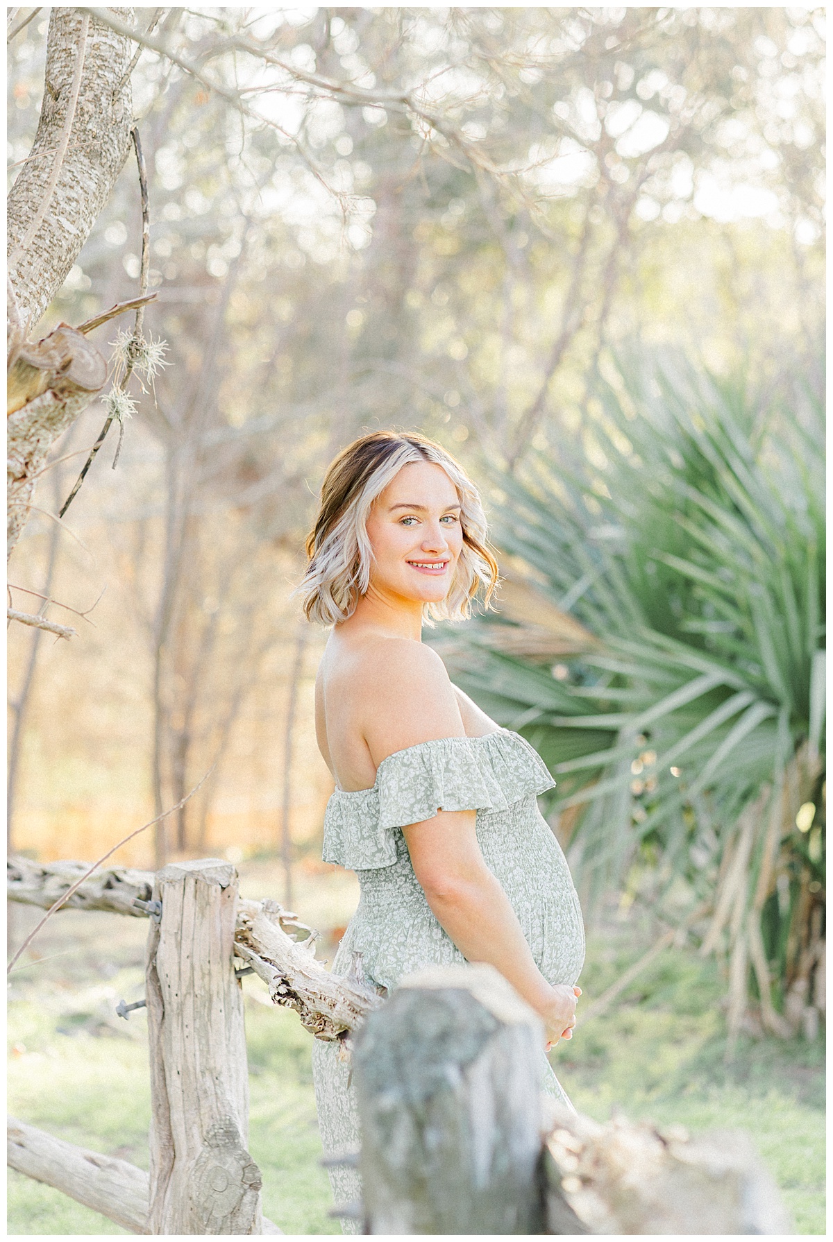 A pregnant mother dressed in a long green dress leans against a rustic fence at Bull Creek District Park. She looks at the camera with a smile during her maternity photoshoot in Austin, TX.