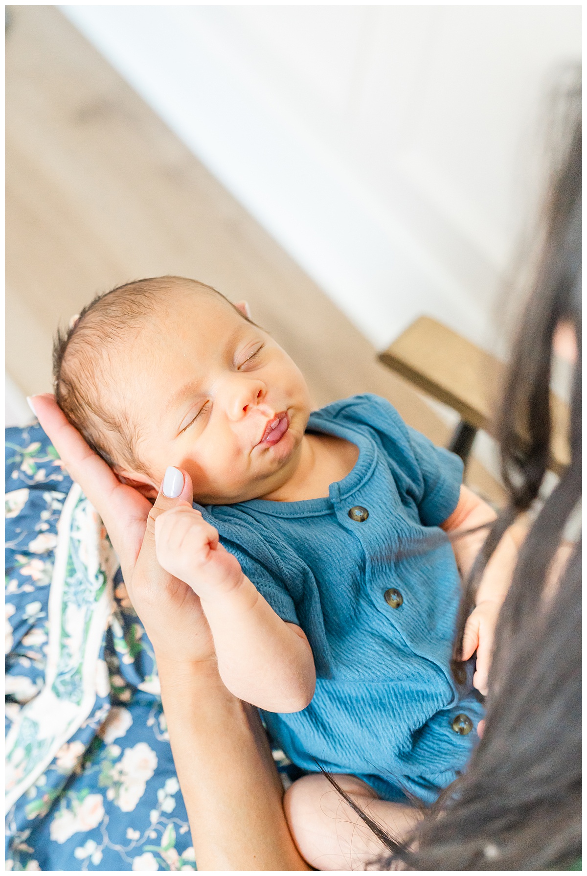 A newborn baby lays on his mother's lap. The mother holds the baby's head in her hand. The baby makes a kiss shape with his mouth while keeping his eyes closed.