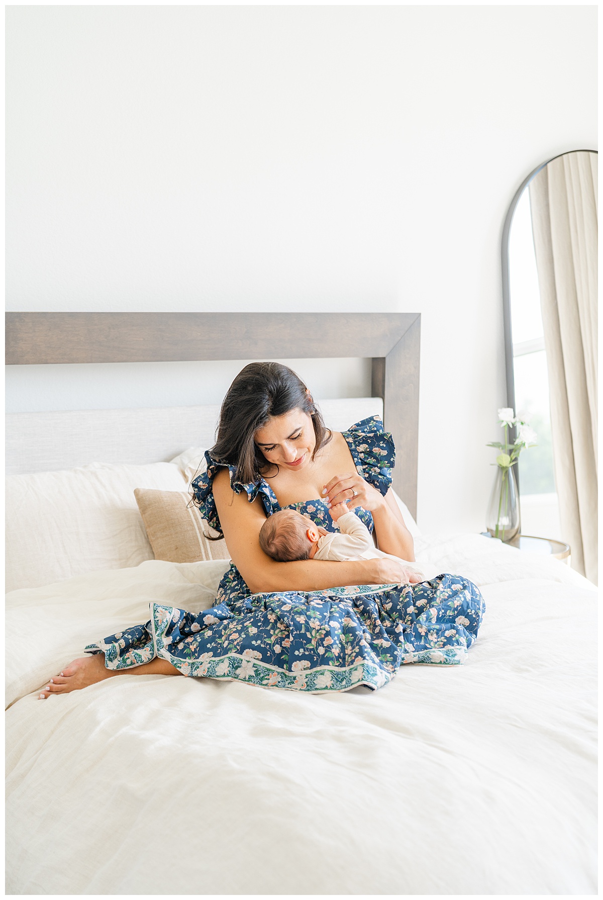 A mother sits on a bed with her newborn baby in her arms. She looks down at her baby with a soft smile, gently captured by a Leander Photographer.