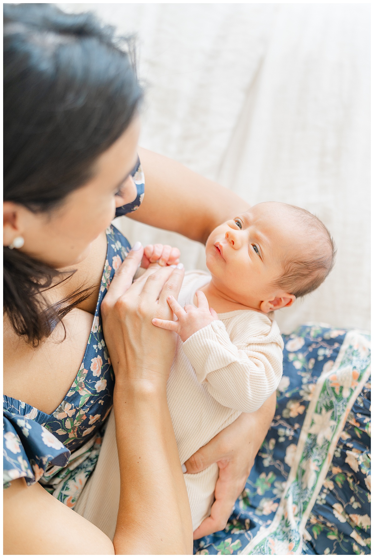 A newborn baby is held by his mother. He holds his mother's fingers with his hand while looking at her, in this candid moment captured by a Leander Photographer.