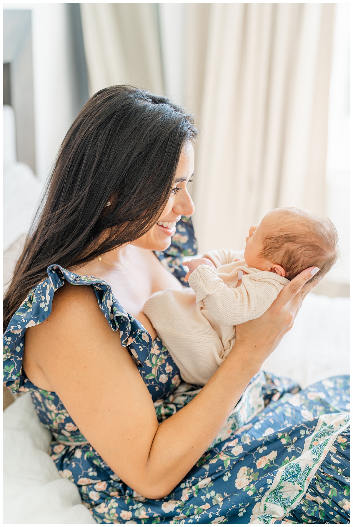 A mother sits on a bed with her newborn baby's bottom sitting against her chest. She looks at her baby with a bright smile on her face.
