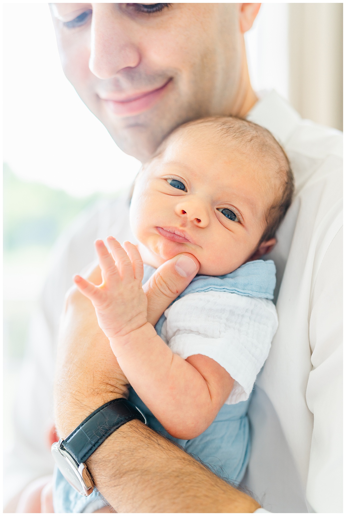 A newborn baby dressed in blue overalls is held by his father while looking at the camera of a Leander Photographer during his in-home newborn session in Leander, TX.