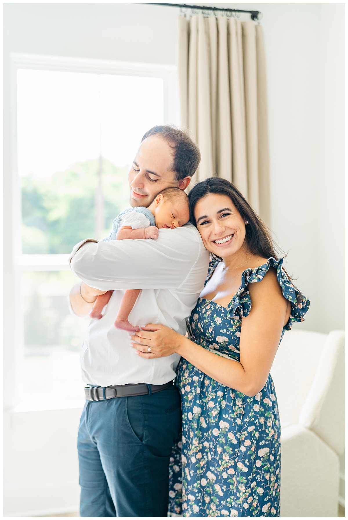 A mother and father stand in front of a window in the Leander home. The father holds their newborn baby up on his shoulder while the mother wraps her arms around her husband.