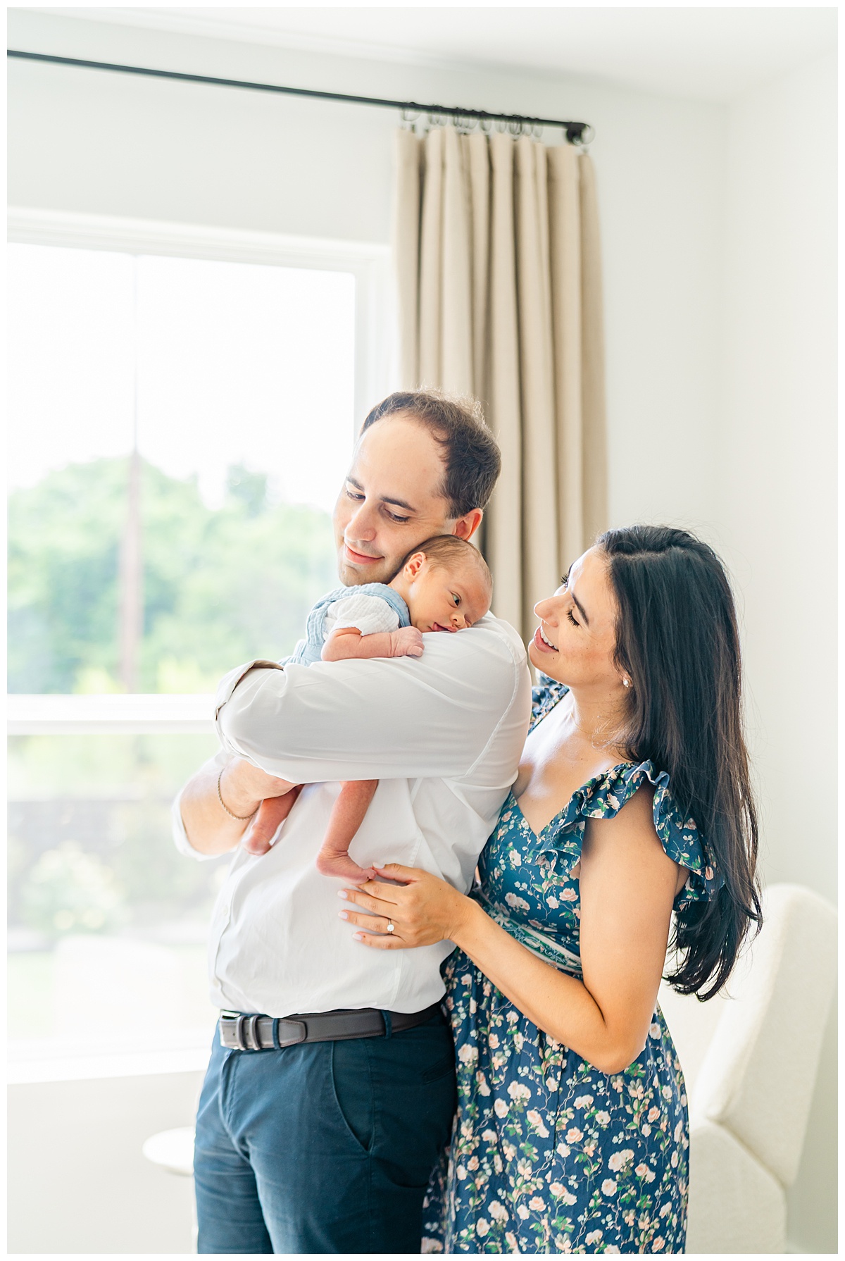 A father holds his newborn baby on his shoulder while he stands in front of a window. The mother stands close behind her husband and looks at her baby while smiling in a candid moment captured by a Leander Photographer.