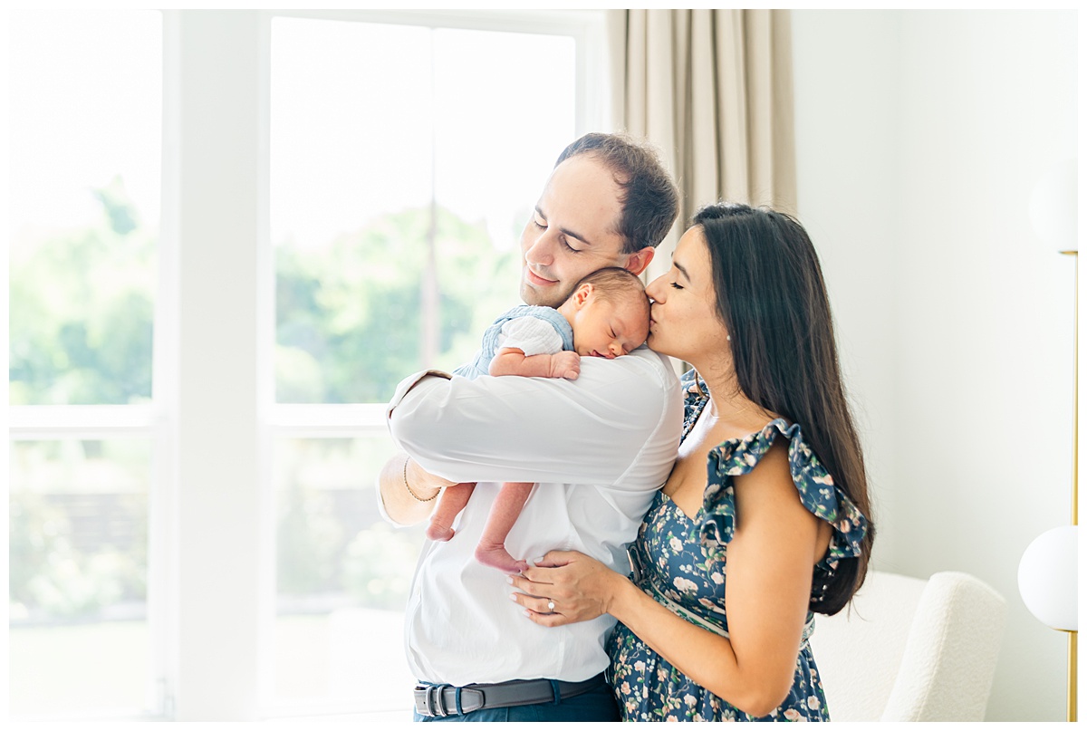 a mother and father stand in front of their bedroom window. The father holds their newborn on his shoulder. The mom stand close behind her husband and kisses their baby on the forehead.