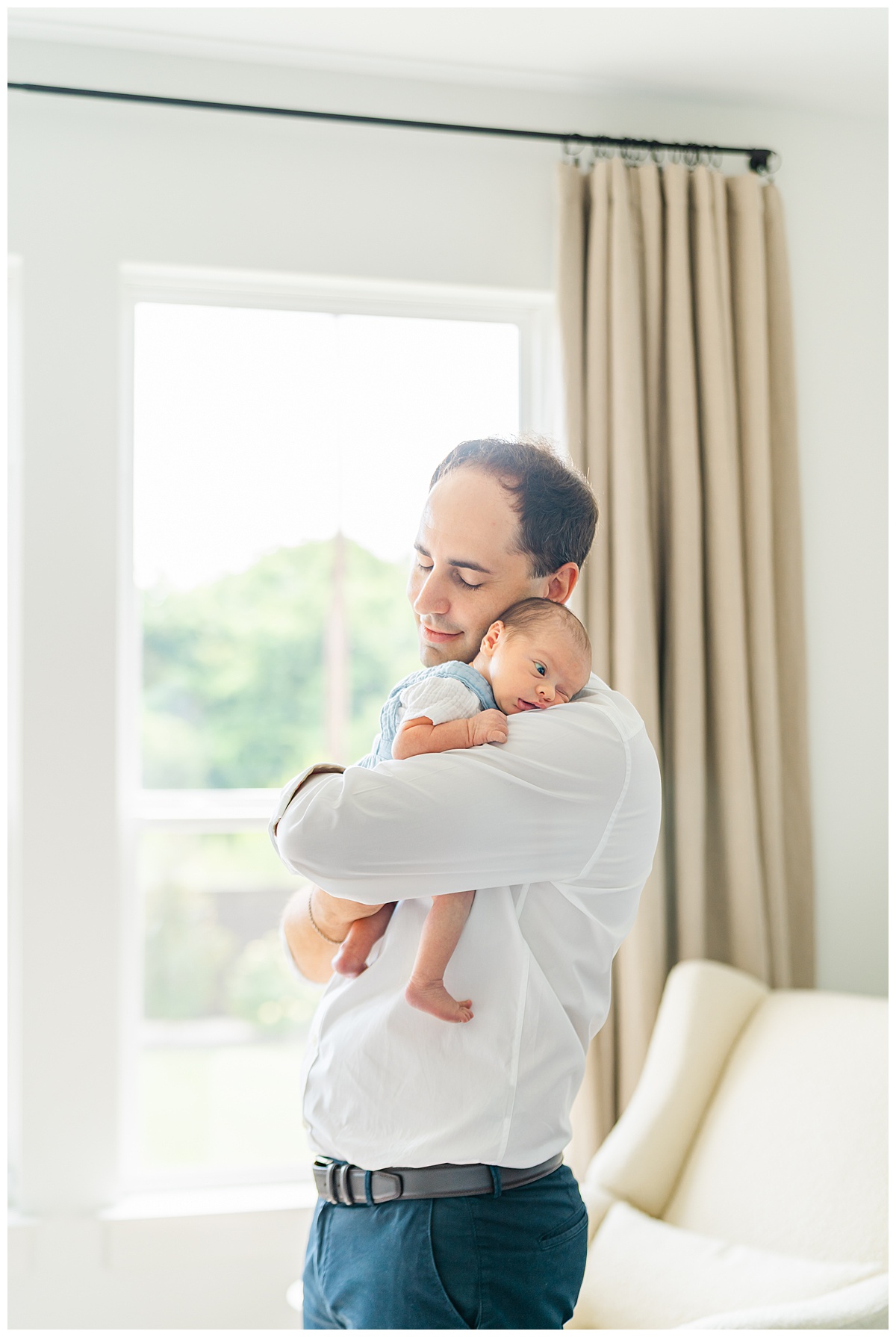 A father stands in front of a window and holds his newborn baby on his shoulder. The baby, dressed is blue overalls, has his eyes opened. The father leans his head against his baby's bed and closes his eyes. 