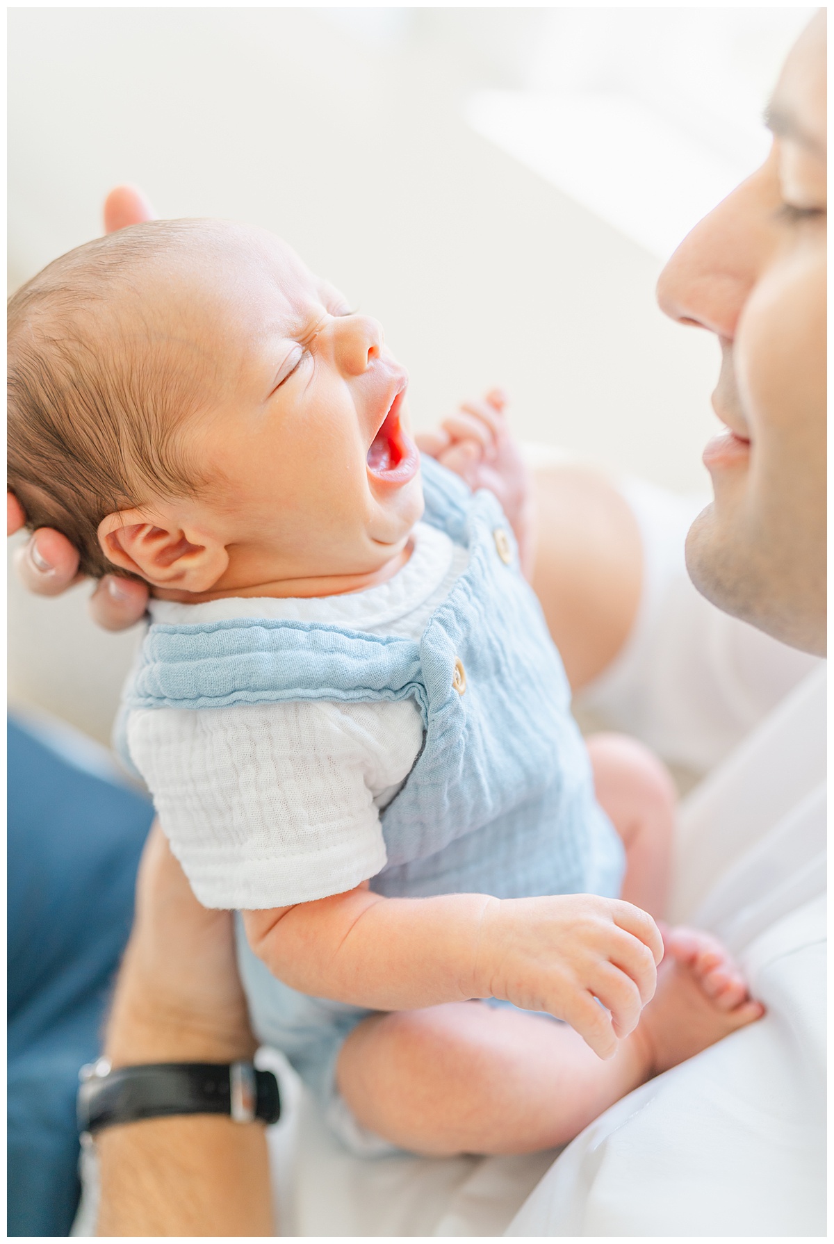 a newborn baby is held by his father and yawns while his father looks down at him in this sweet shot captured by a Leander Photographer.