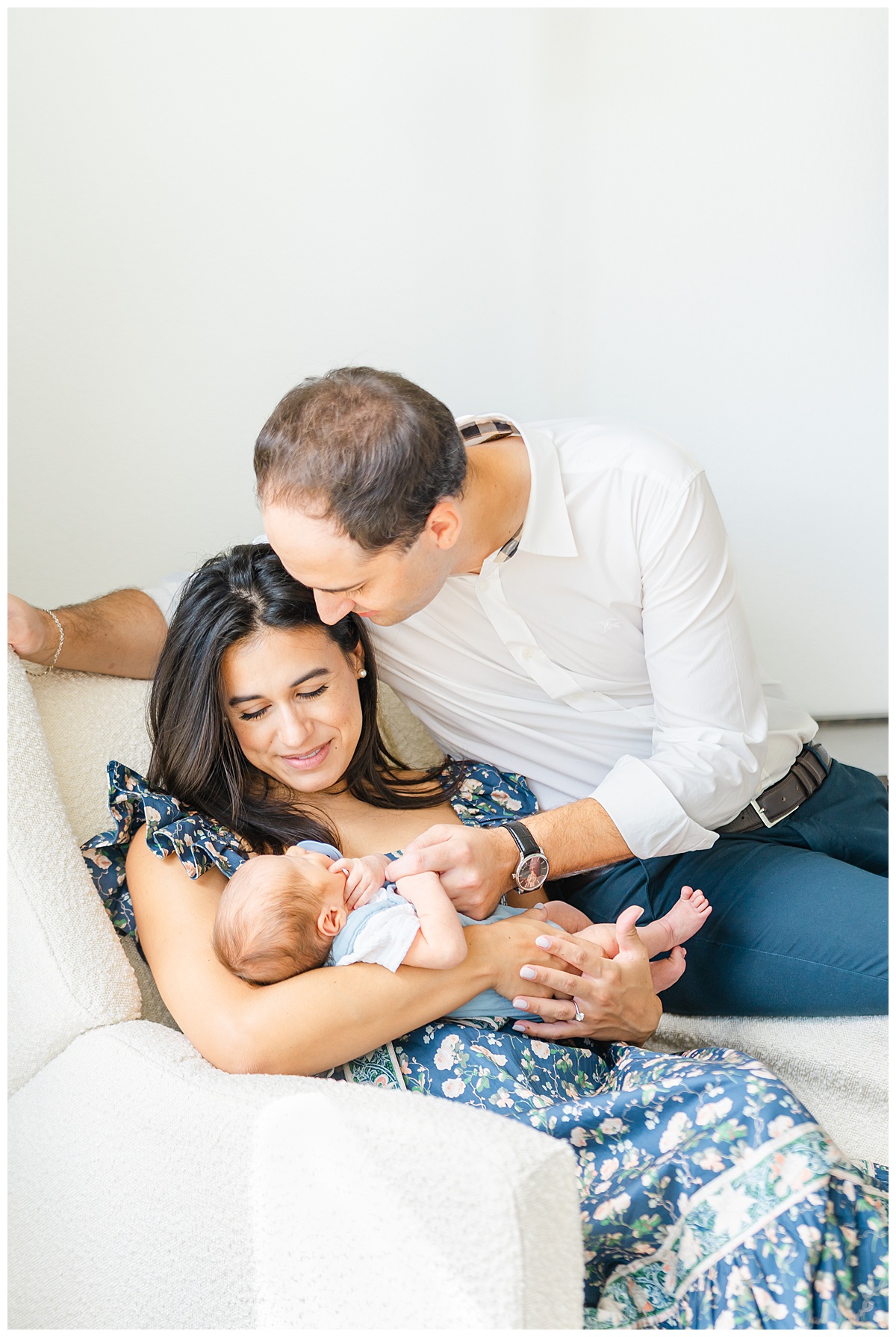 A mother sits in a rocking chair holding her newborn baby in their Leander home. Her husband sits on the side of the chair. They both look down at their baby with a smile.