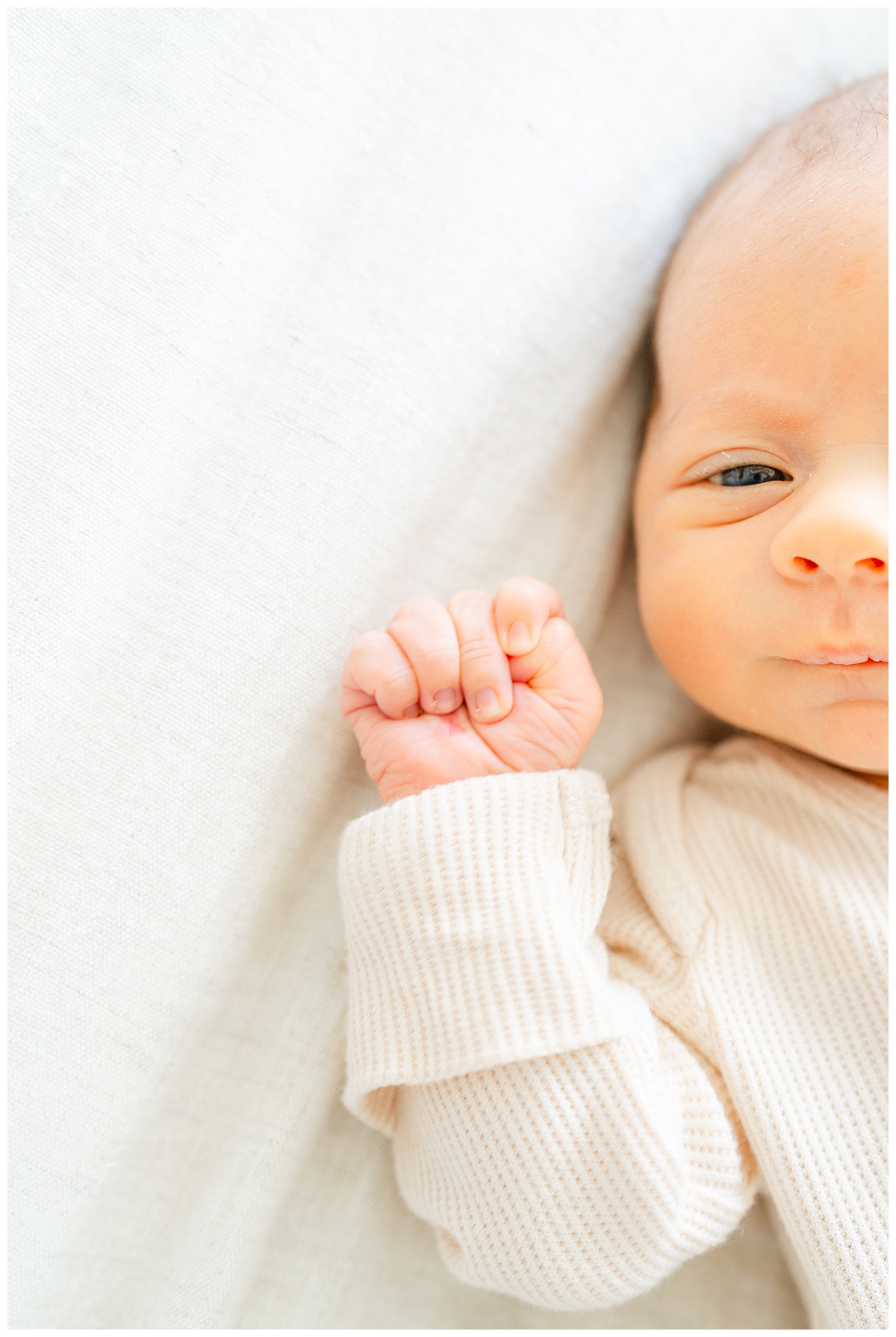 Close of photo of a newborn baby's hand. The baby has his eyes open and lays on a beige comforter.