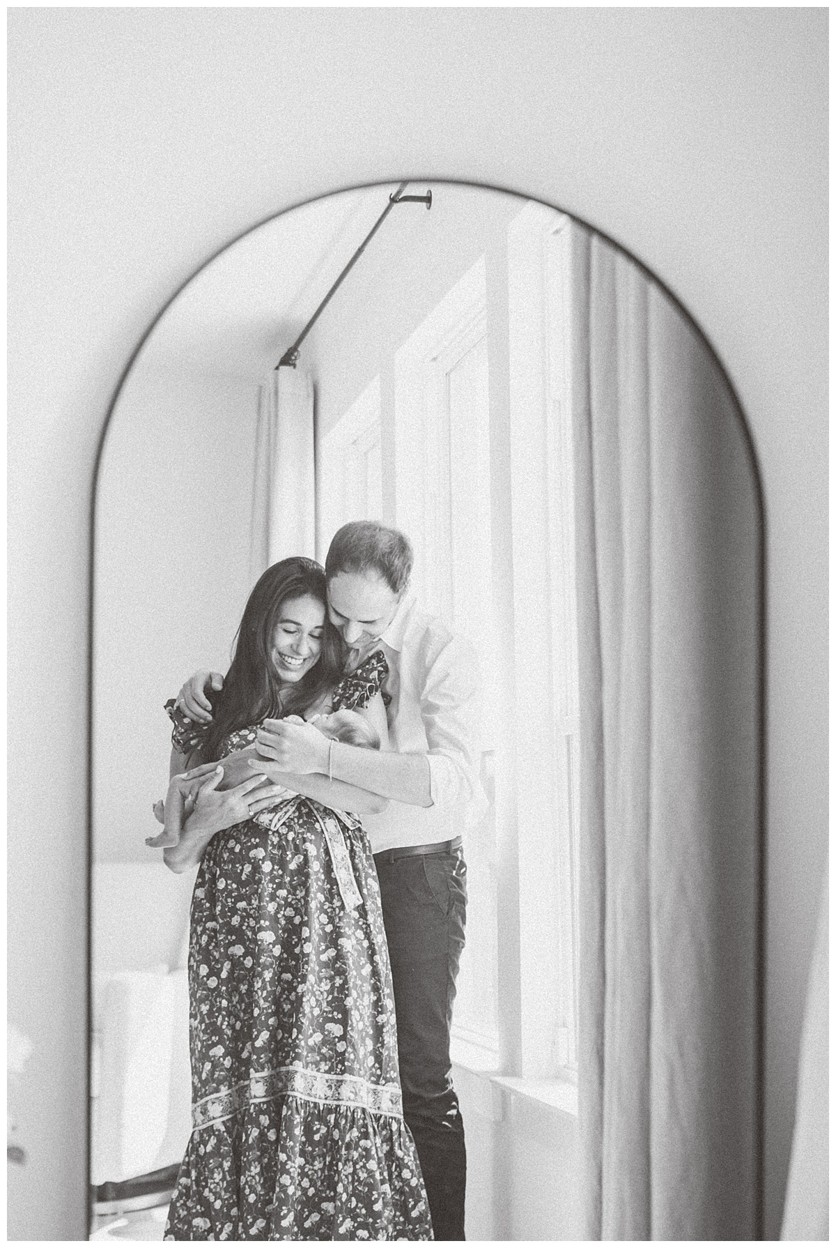 A mother and father stand in front of a tall mirror. The mother holds their newborn baby. They are both looking at their baby with a smile. The shot is captured through the mirror by a Leander Photographer.