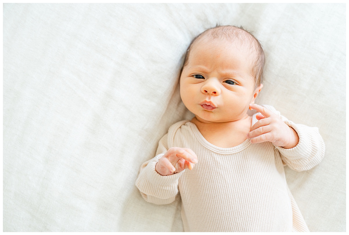 a newborn baby lays on a bed. He makes a kiss shape with his mouth while looking at the camera.