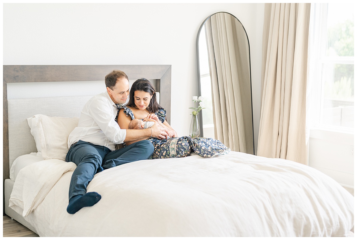A father and mother sit on their bed in their Leander home. The mother holds their newborn baby. They both look down at their baby with a smile.
